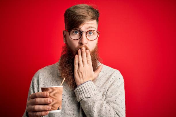 istock Irish redhead man with beard drinking a takeaway coffee on a paper cup over red background cover mouth with hand shocked with shame for mistake, expression of fear, scared in silence, secret concept 1204794482