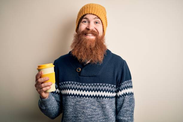 Redhead Irish man with beard wearing winter hat drinking take away hot coffee with a happy face standing and smiling with a confident smile showing teeth