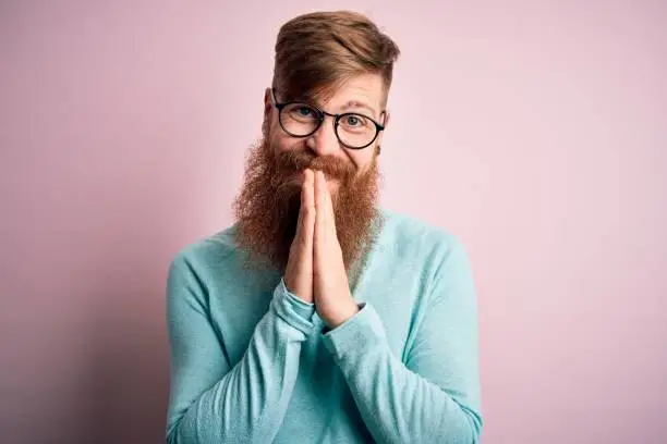 Handsome Irish redhead man with beard wearing glasses over pink isolated background praying with hands together asking for forgiveness smiling confident.