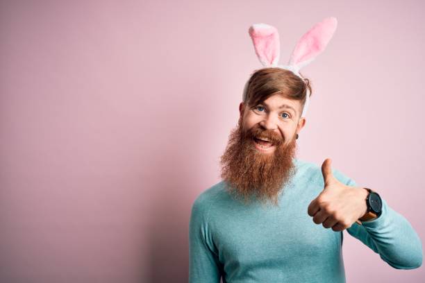 istock Hipster Irish man with beard wearing easter rabbit ears over isolated pink background doing happy thumbs up gesture with hand. Approving expression looking at the camera showing success. 1204794761
