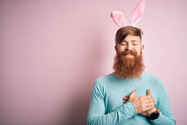 istock Hipster Irish man with beard wearing easter rabbit ears over isolated pink background smiling with hands on chest with closed eyes and grateful gesture on face. Health concept. 1204794764