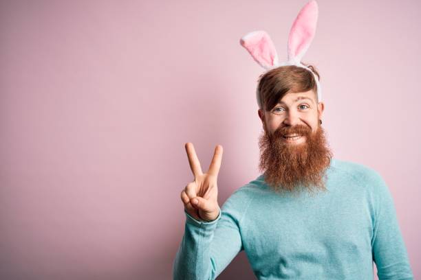istock Hipster Irish man with beard wearing easter rabbit ears over isolated pink background smiling with happy face winking at the camera doing victory sign. Number two. 1204794769
