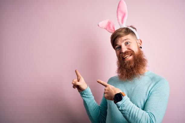 istock Hipster Irish man with beard wearing easter rabbit ears over isolated pink background smiling and looking at the camera pointing with two hands and fingers to the side. 1204794771
