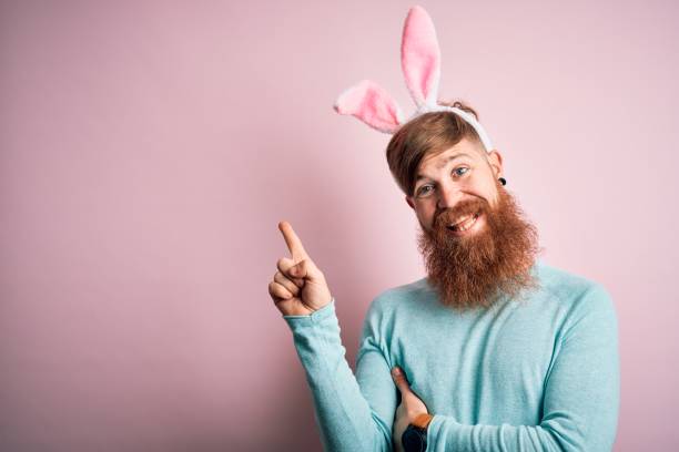 istock Hipster Irish man with beard wearing easter rabbit ears over isolated pink background with a big smile on face, pointing with hand and finger to the side looking at the camera. 1204794774