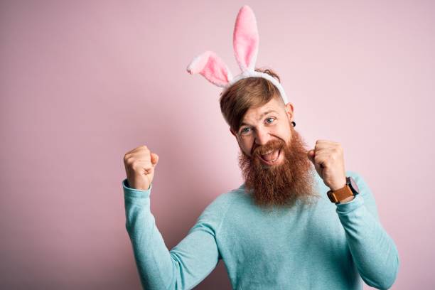 istock Hipster Irish man with beard wearing easter rabbit ears over isolated pink background celebrating surprised and amazed for success with arms raised and open eyes. Winner concept. 1204794775