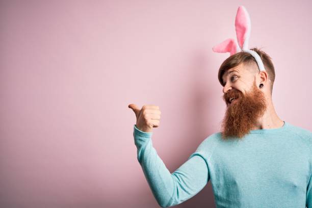 istock Hipster Irish man with beard wearing easter rabbit ears over isolated pink background smiling with happy face looking and pointing to the side with thumb up. 1204794787
