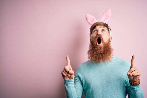 istock Hipster Irish man with beard wearing easter rabbit ears over isolated pink background amazed and surprised looking up and pointing with fingers and raised arms. 1204794793
