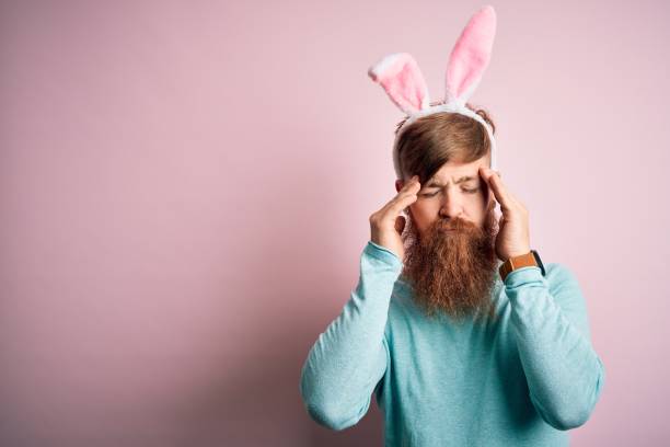 수염을 입은 힙스터 아일랜드 남자는 고립 된 분홍색 배경 위에 부활절 토끼 귀를 입고 두통이 절실하고 통증과 편두통 때문에 스트레스를 받았습니다. 머리에 손을. - irish culture audio 뉴스 사진 이미지