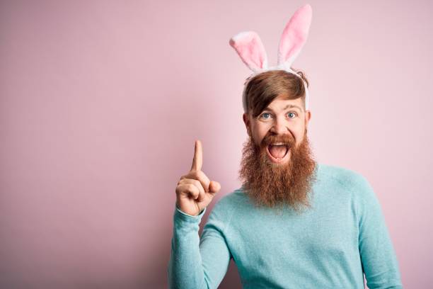 istock Hipster Irish man with beard wearing easter rabbit ears over isolated pink background pointing finger up with successful idea. Exited and happy. Number one. 1204794803