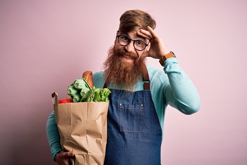 Redhead Irish supermarket worker man with beard wearing apron and holding fresh groceries stressed with hand on head, shocked with shame and surprise face, angry and frustrated. Fear and upset for mistake.