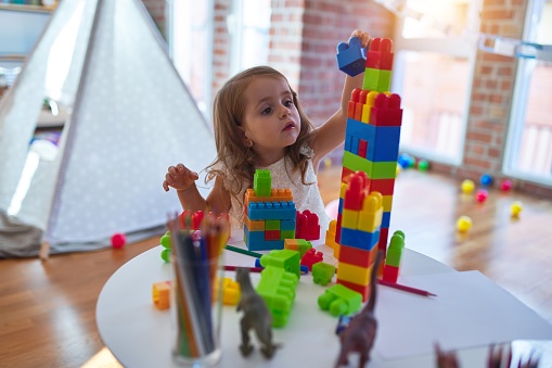 Adorable blonde toddler playing with building blocks around lots of toys at kindergarten