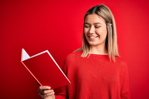 Young beautiful woman reading book standing over isolated red background with a happy face standing and smiling with a confident smile showing teeth