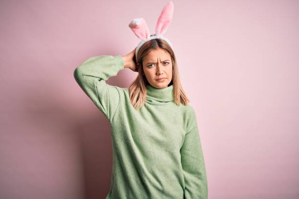Young beautiful woman wearing easter rabbit ears standing over isolated pink background confuse and wonder about question. Uncertain with doubt, thinking with hand on head. Pensive concept.