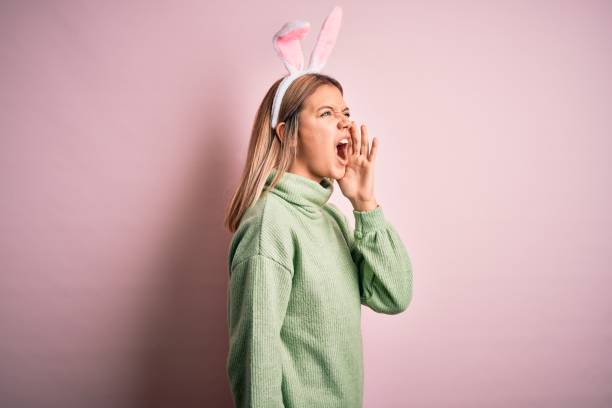 jeune belle femme utilisant des oreilles de lapin de pâques restant au-dessus du fond rose d’isolement criant et criant fort à côté avec la main sur la bouche. concept de communication. - easter rabbit bizarre women photos et images de collection