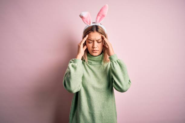 istock Young beautiful woman wearing easter rabbit ears standing over isolated pink background with hand on head for pain in head because stress. Suffering migraine. 1204798264