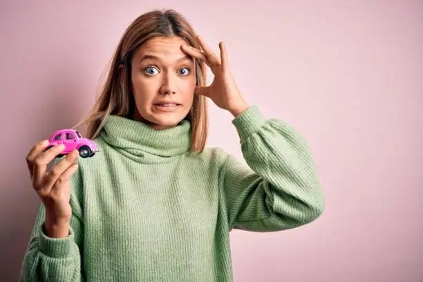 Photo of Young beautiful woman holding small toy car standing over isolated pink background stressed with hand on head, shocked with shame and surprise face, angry and frustrated. Fear and upset for mistake.