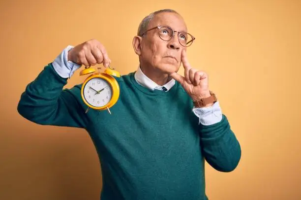 Photo of Senior handsome man holding alarm clock standing over isolated yellow background serious face thinking about question, very confused idea