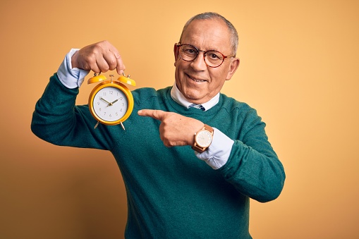 Senior handsome man holding alarm clock standing over isolated yellow background very happy pointing with hand and finger