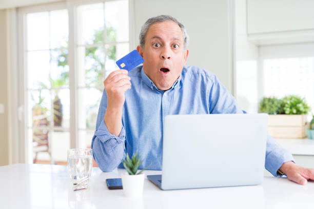 집에서 신용 카드와 노트북을 사용하여 온라인으로 쇼핑하는 잘 생긴 노인은 깜짝 얼굴로 충격에 무서워하고 두려움과 두려움 표현으로 흥분했습니다. - mouth open retail shock surprise 뉴스 사진 이미지