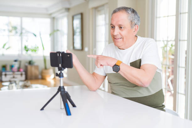 istock Handsome senior man doing video conference talking to the smartphone camera amazed and smiling to the camera while presenting with hand and pointing with finger. 1204805333