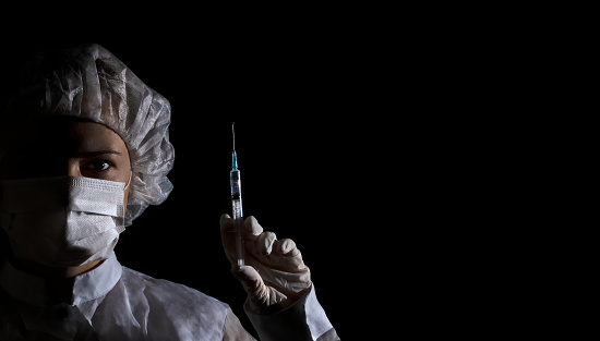 Female doctor in protection clothing holds syringe with vaccine in her hand on black background. Half face in shadow, dramatic emotion. Concept of virus protection, prevention of epidemics, treatment of diseases