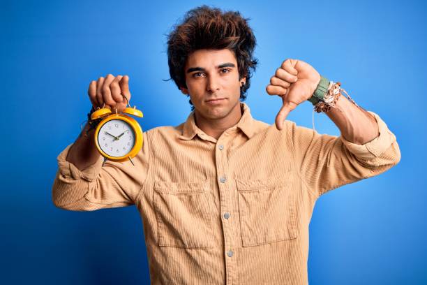 istock Young handsome man holding alarm clock standing over isolated blue background with angry face, negative sign showing dislike with thumbs down, rejection concept 1204810417
