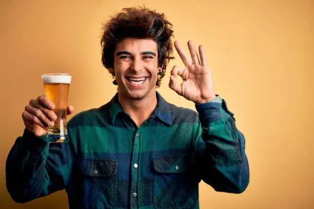 Photo of Young handsome man drinking glass of beer standing over isolated yellow background doing ok sign with fingers, excellent symbol