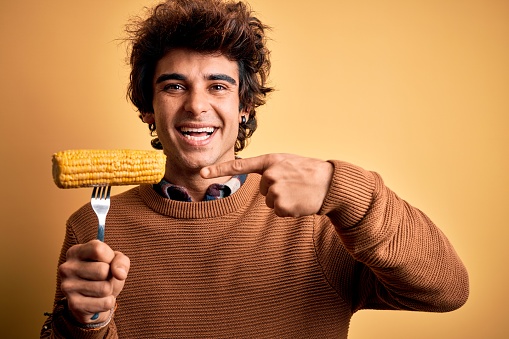 Young handsome man holding fork with cob corn standing over isolated yellow background very happy pointing with hand and finger