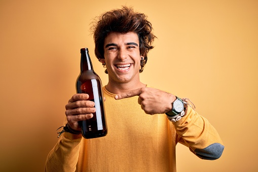 Young handsome man drinking bottle of beer standing over isolated yellow background very happy pointing with hand and finger