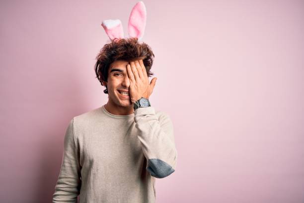 istock Young handsome man holding easter rabbit ears standing over isolated pink background covering one eye with hand, confident smile on face and surprise emotion. 1204811384