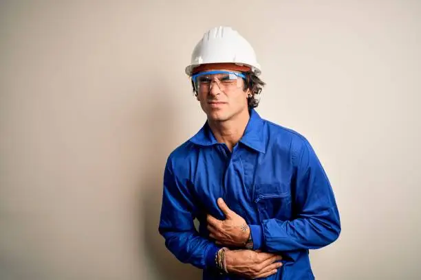 Photo of Young constructor man wearing uniform and security helmet over isolated white background with hand on stomach because nausea, painful disease feeling unwell. Ache concept.