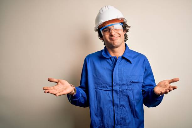 istock Young constructor man wearing uniform and security helmet over isolated white background clueless and confused expression with arms and hands raised. Doubt concept. 1204811571