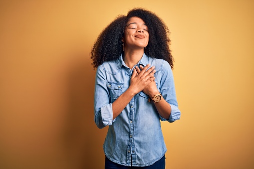 Young beautiful african american woman with afro hair standing over yellow isolated background smiling with hands on chest with closed eyes and grateful gesture on face. Health concept.
