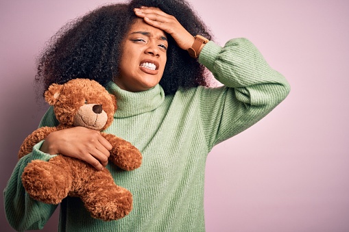 Young african american woman with afro hair hugging teddy bear over pink background stressed with hand on head, shocked with shame and surprise face, angry and frustrated. Fear and upset for mistake.