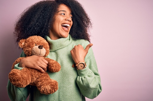 Young african american woman with afro hair hugging teddy bear over pink background pointing and showing with thumb up to the side with happy face smiling