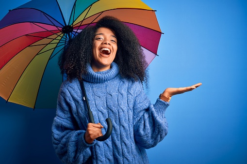 Young african american woman with afro hair under colorful umbrella for winter weather rain very happy and excited, winner expression celebrating victory screaming with big smile and raised hands
