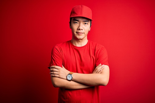Young handsome chinese delivery man wearing cap standing over isolated red background skeptic and nervous, disapproving expression on face with crossed arms. Negative person.