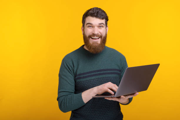 istock Photo of happy young man using laptop while standing over yellow background 1204839580