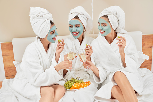 Front view of group of girls with cosmetic masks, in towels and bathrobes having home party. Three pretty female friends clinking glasses with wine, talking, laughing and eating grapes in bedroom.
