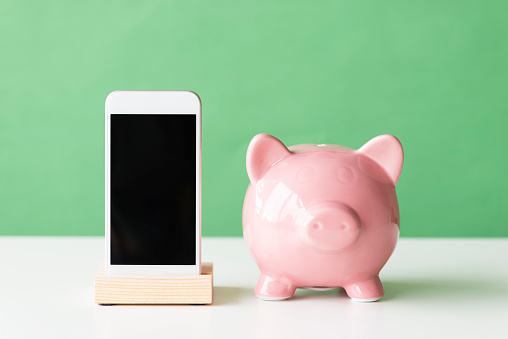 Front view of a smart phone and pink piggy bank on white table in front of a green wall.