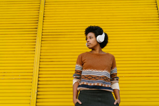 Young woman listening to music A young black woman is listening to music with a bluetooth wireless headphones connected to her portable music player in front of a yellow wall. in front of photos stock pictures, royalty-free photos & images