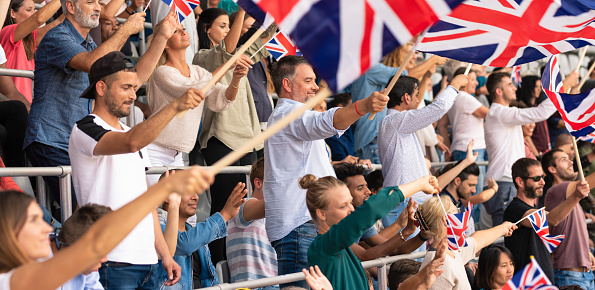 Large crowd on a stadium cheering for their team with their hands raised and waving Great Britain flags.