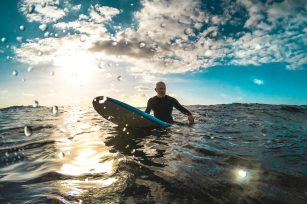 surfer entspannen auf surfbrett bei sonnenuntergang auf teneriffa warten auf die nächste gute welle - sport reisekonzept mit flacher schärfentiefe mit tropfen auf der linse als zusammensetzung - kontrast halo sonnenuntergang filter - surfing men hawaii islands wave stock-fotos und bilder