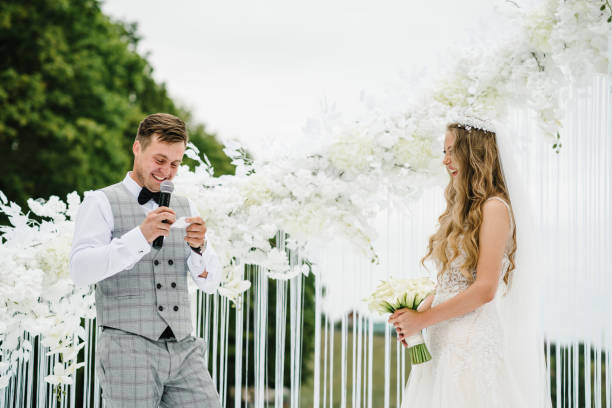 istock Happy bride and groom exchanging wedding vows on ceremony under the arch decorated with flowers and greenery on the backyard banquet area. The groom says the promise in the microphone. 1204862642