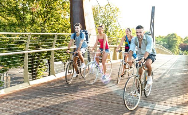 amigos milenarios felices divirtiéndose montando en bicicleta en el parque de la ciudad - concepto de amistad con jóvenes estudiantes milenarios andando juntos en carril bici - filtro brillante por la tarde con halo de sol - late spring fotografías e imágenes de stock