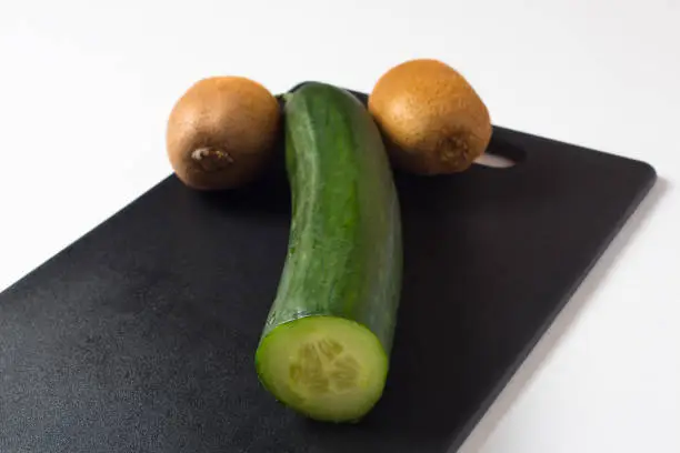 A cucumber and two kiwis lie on a black cutting Board. Diet food and salads for a healthy lifestyle. The likeness of a phallic symbol or masculine dignity. Fresh vegetables and fruits.