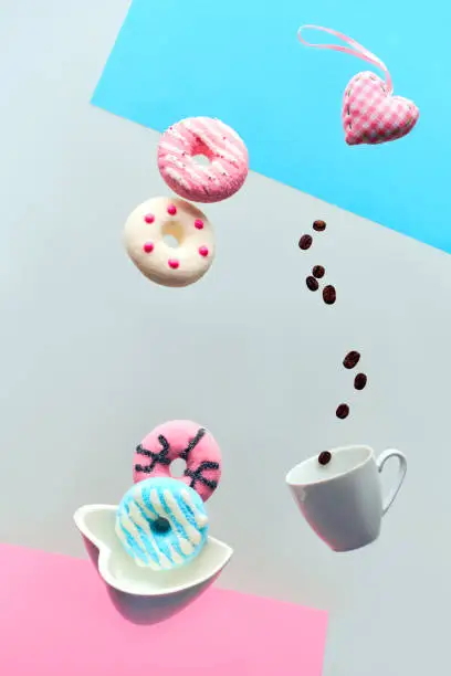 St. Valentine concept, levitation of doughnuts above heart shaped bowl. Coffee beans flying in espresso cup. Hand catching pink doughnut. Creative background in pink, blue mint and light grey color.