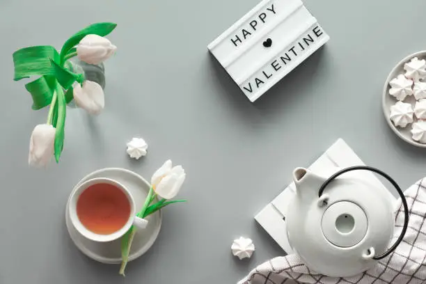 Geometric panoramic flat lay with hands. Cup of black tea, tea pot, sweets and white tulips on grey with lightboard. Text Happy Valentine day on the light board. Geometric trendy top view.