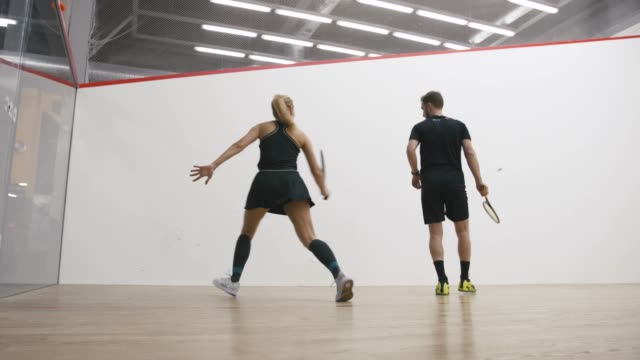 Young athletic man and woman play squash together in the squash court, slow motion, low angle view