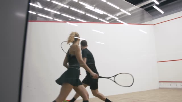 Young athletic man and woman play squash together in the squash court, slow motion, camera is behind the glass